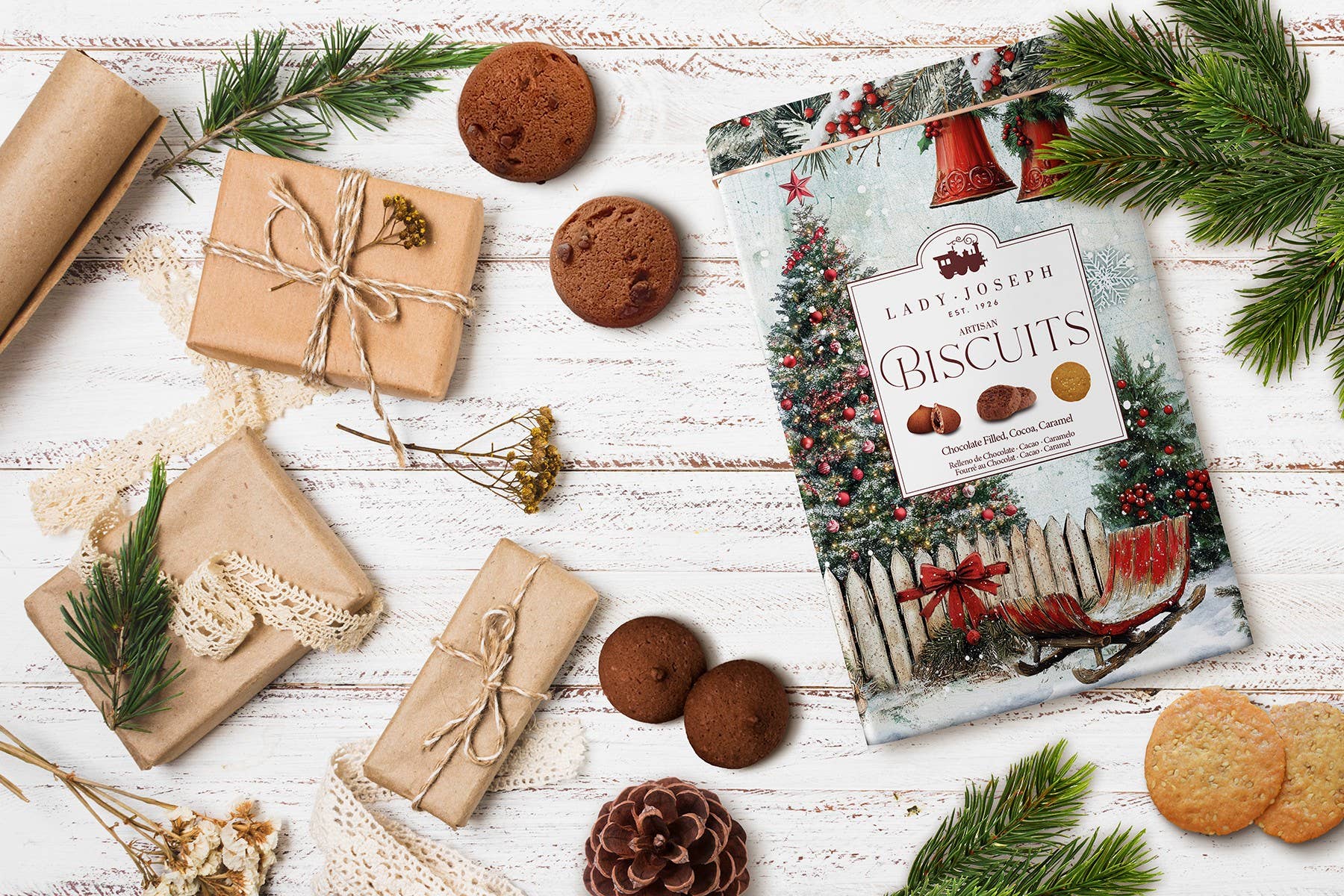 Ein festliches Flat Lay zeigt die Lady Joseph - Sortiment von Keksen in einer Metallgeschenkbox für Weihnachten, eingepackte Geschenke, Tannenzapfen, Grünzeug und gemischte Kekse auf einer weißen Holzoberfläche.