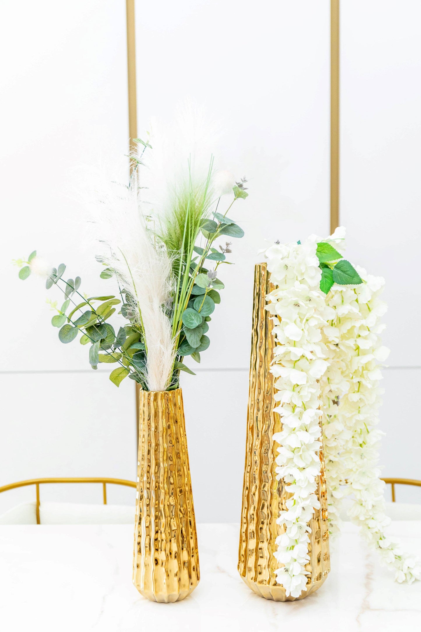 Zwei hohe AULICA – GLÄNZENDE GOLDENE VASE (15,5 x 15,5 x 55,5 cm) stehen auf einem weißen Untergrund. Eine Vase enthält grünes Laub, Pampasgras und Eukalyptus; die andere präsentiert kaskadenförmige weiße Blüten. Der weiß-goldene Hintergrund unterstreicht ihre Eleganz.