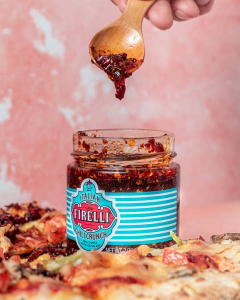 Eine Hand schöpft 200 g Casa Firelli Chili Crunch aus einem Firelli-Glas, hergestellt mit italienischem Olivenöl und kalabrischen Chiliflocken, mit Pizza belegt mit Chili Crunch vor einem rosa Hintergrund.