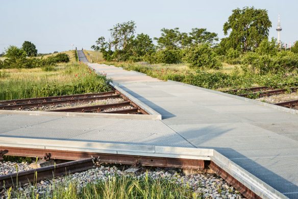 Ein Metallsteg aus der Landschaftsarchitektur 2025 des Callwey Verlags wölbt sich über alte Bahngleise in einem grasbewachsenen Gebiet, in dessen Hintergrund ein hoher Fernmeldemast zu sehen ist.