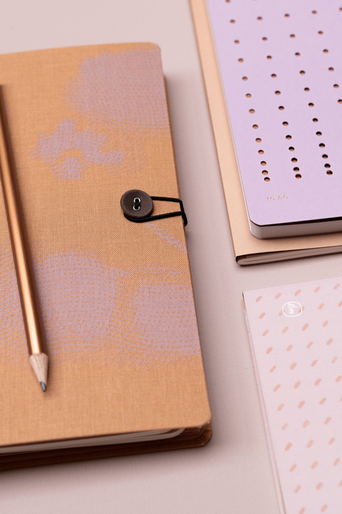 Eine Nahaufnahme der Tinne Mia Aufgabenliste in Lavendel, mit Button-Verschluss, goldfarbenem Stift und kleinem nachhaltig produziertem Notizblock, alles ordentlich auf einer hellen Fläche arrangiert.