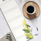 Eine Tasse schwarzen Kaffees auf einer hölzernen Untertasse ruht neben dem Wildblumen Atelier - Wandkalender 2026 mit handgezeichneten Blumen (FSC), der bis März geöffnet ist, geschmückt mit gelben Mimosenblüten auf einer Marmorfläche mit Olivenblättern und einer beigen Serviette.