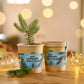 Zwei biologisch abbaubare Stadtgärtner-Tassen mit festlichen Etiketten stehen auf einem Holztisch. In einem steht das Anzuchtset - Weihnachtsbaum aus dem Becher. Dahinter verschwimmen warme Lichterketten und goldenes Dekor und schaffen eine gemütliche Weihnachtsstimmung - ideal für ein Weihnachtsgeschenk.