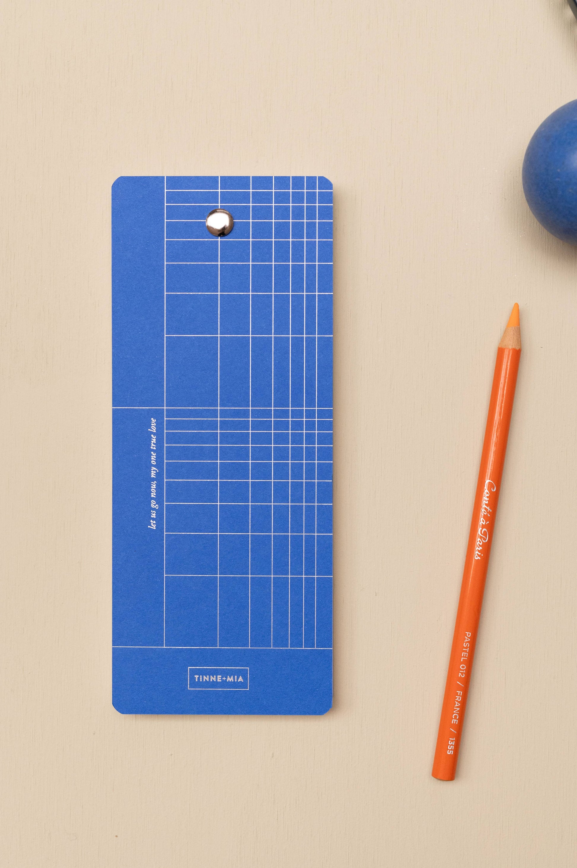 Die Tinne Mia Aufgabenliste "True Blue" aus nachhaltigem, rechteckigem FSC-Papier mit weißen Linien und Metallring liegt neben einem orangenen Bleistift auf Beige; in der Ecke befindet sich eine dunkelblaue Kugel.