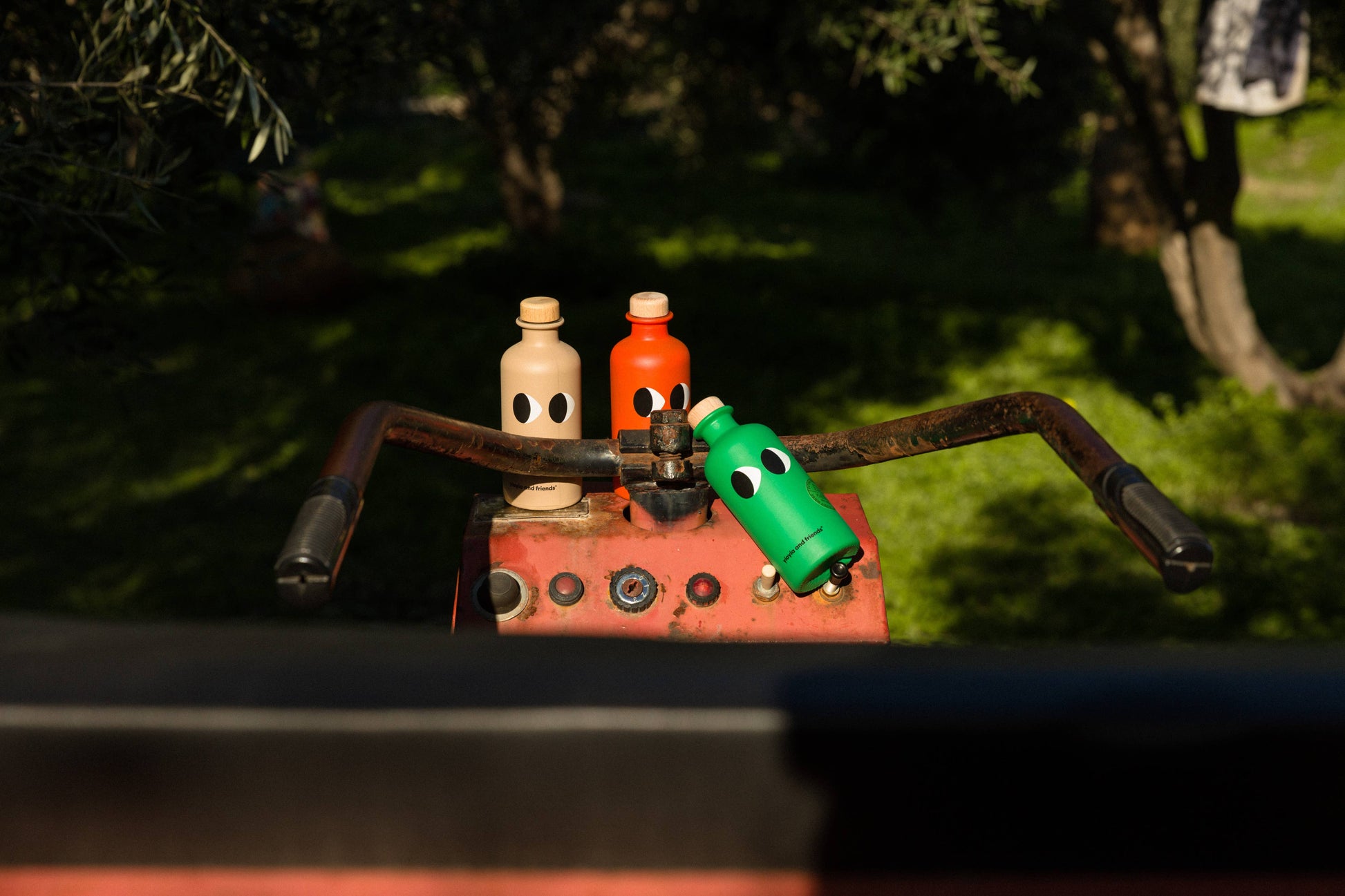 Drei leuchtende yiayia and friends-Flaschen mit verspielten Gesichtern sitzen auf roten Lenkern im Freien. Das Natives Olivenöl extra mit Basilikum von yiayia and friends hebt sich von grünem Gras und sonnigen Bäumen ab.