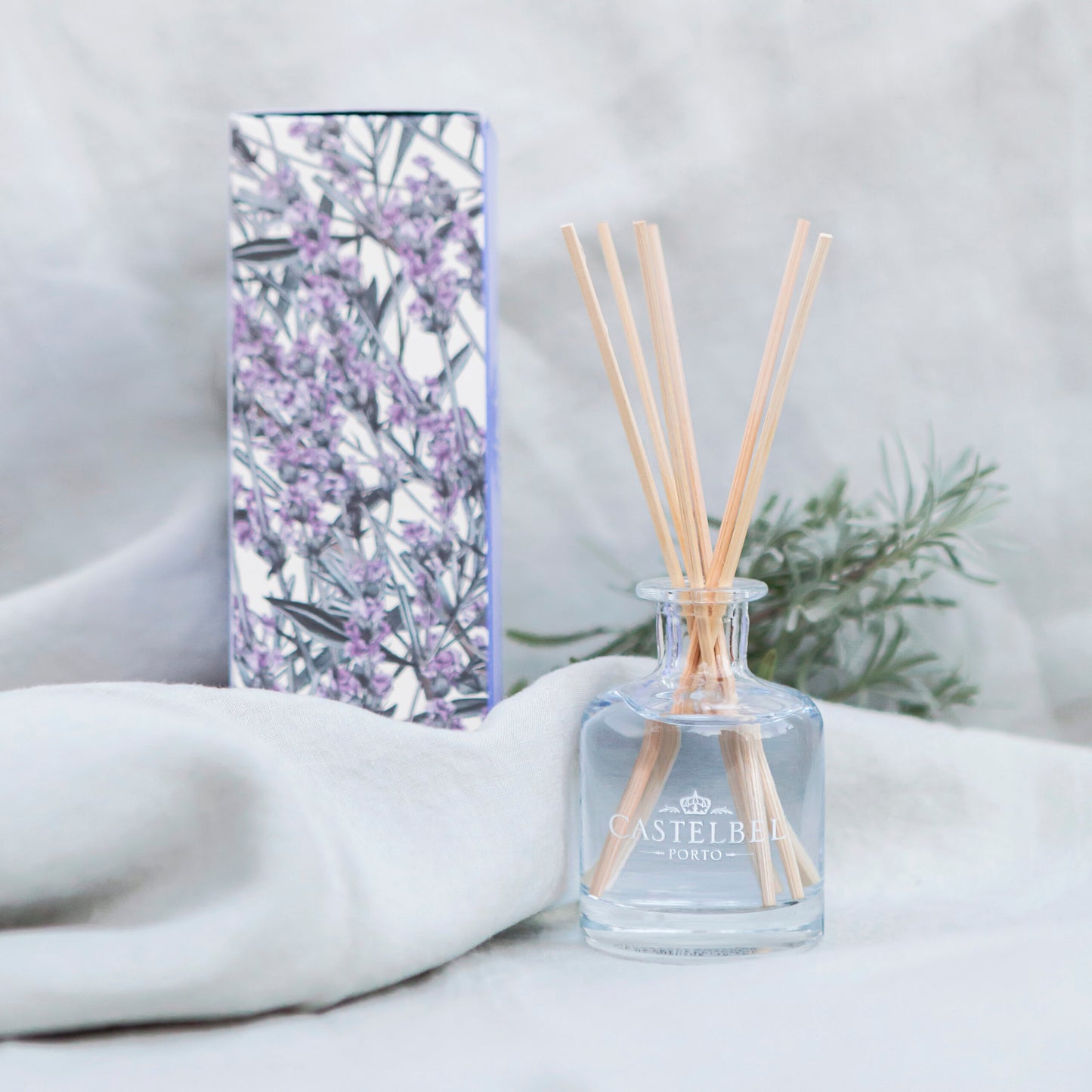 Eine Glasflasche Castelbel Diffusor | Lavendel 100 ml mit Holzstäbchen steht auf einem weichen, weißen Stoff, daneben ein Blumenkasten und Lavendelzweige im Hintergrund.