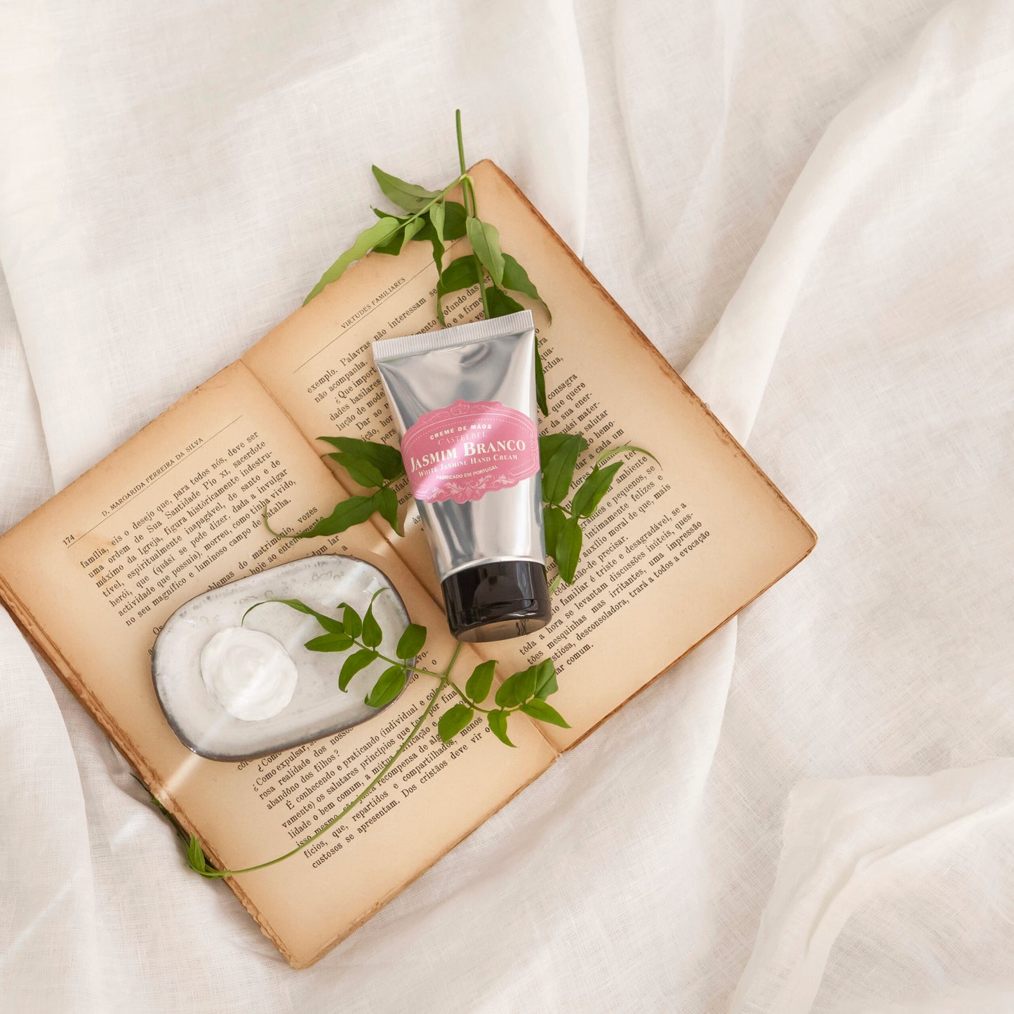Eine silberne Tube Castelbel Handcreme | Jasmin 60 ml mit rosafarbenem Etikett liegt neben einer Seifenschale auf einem aufgeschlagenen Buch, umgeben von grünen Blättern, die an natürliche Jasminfrische und intensive Feuchtigkeitspflege erinnern.