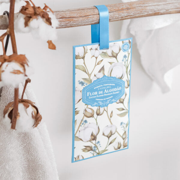 Ein Castelbel Sachet | Cotton Flower 10 g hängt an einem blauen Band an einem Holzstab neben Baumwollzweigen und einem weißen Handtuch. Auf der Vorderseite ist ein Blumenmotiv mit portugiesischem Text abgebildet.