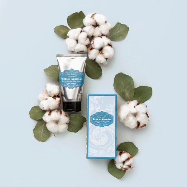 Eine Castelbel Handcreme | Cotton 60ml Tube und die dazugehörige Schachtel sind auf einer hellblauen Fläche ausgestellt, umgeben von Baumwollzweigen mit grünen Blättern und flauschigen weißen Baumwollkapseln.