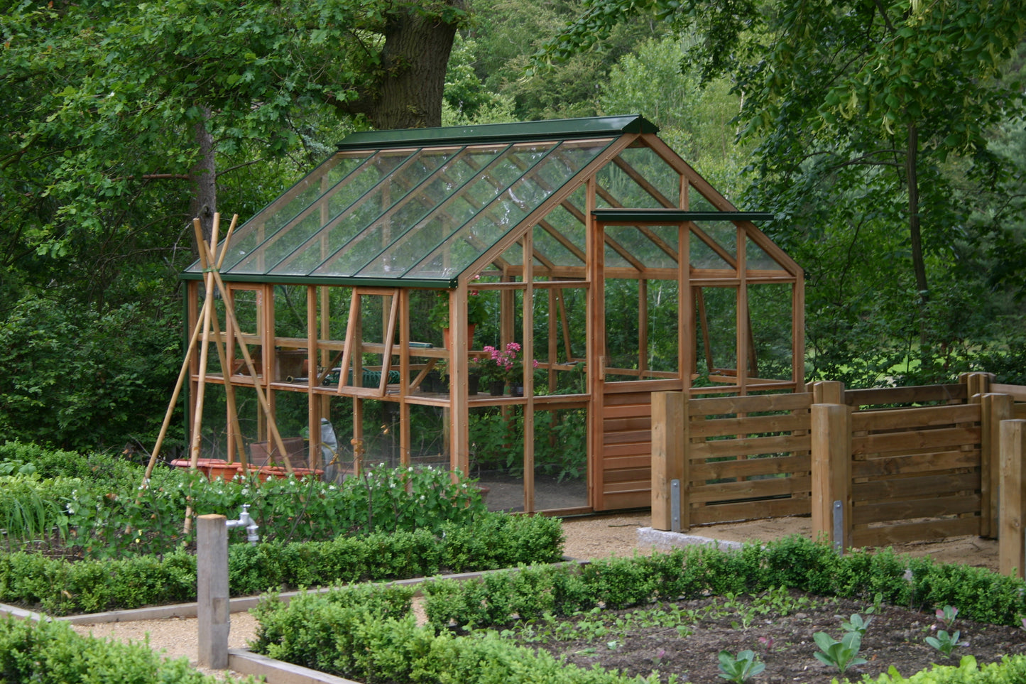 In einem üppigen Garten findet der Workshop "Aus eigener Anzucht: selbst Pflanzen vermehren" von Dr. Cathrine Thorpe am Sa. 21.03.2026 (14:00-16:00) inmitten von Grünpflanzen und Gemüse, die in Hochbeeten neben einem Glasgewächshaus mit Holzrahmen gedeihen.