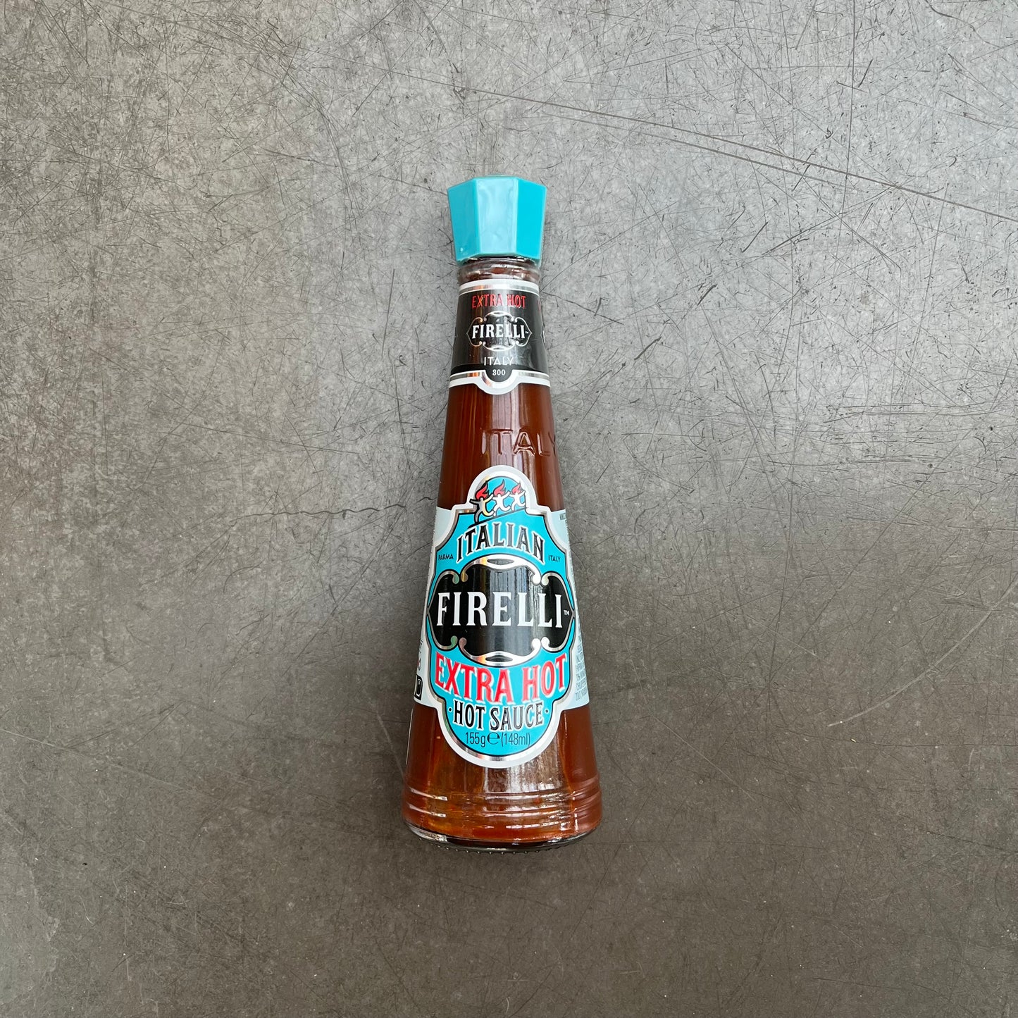 A 148ml Glasflasche Firelli - Casa Firelli Extra scharfe italienische Sauce von Firelli mit blauem Deckel und Etikett steht aufrecht auf einer strukturierten grauen Oberfläche.