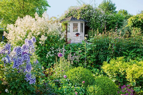 Ein kleiner weißer Gartenpavillon mit Tisch und Stuhl ist umgeben von üppigen Grünpflanzen, bunt blühenden Stauden im Garten aus dem Callwey Verlag und kreativen Blumenmustern in einem lebendigen, sonnenbeschienenen Garten.