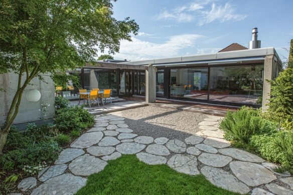 Modernes Haus mit großen Glaswänden, Steinterrasse und Garten. Essplatz im Freien mit gelben Stühlen im Grünen - ein inspirierendes Beispiel aus "Landschaftsarchitektur 2025" des Callwey Verlags, das moderne Landschaftsgestaltung unter blauem Himmel zeigt.