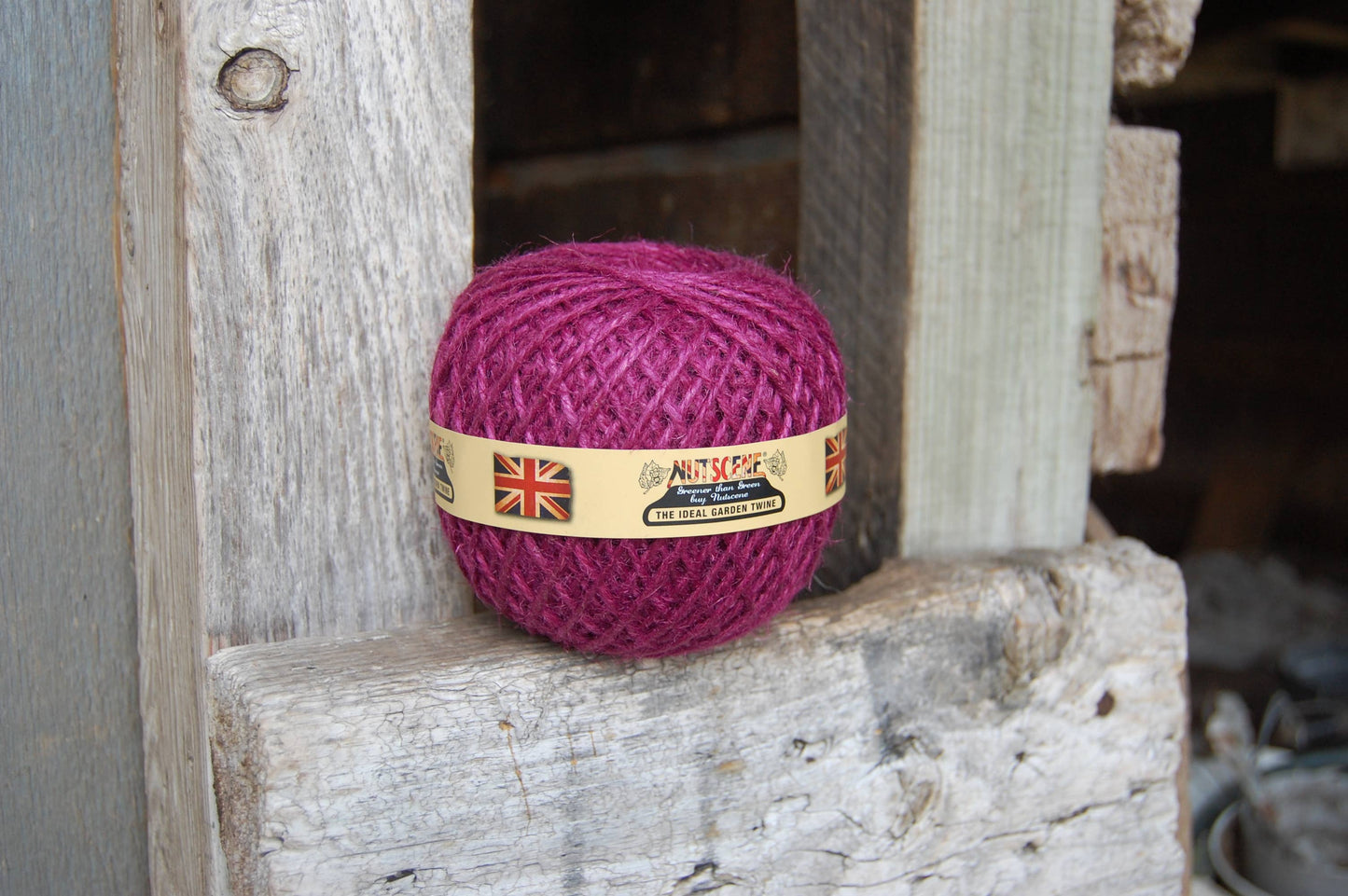 Ein magentafarbenes Knäuel des Heritage Twine von Nutscene Twines Ltd. sitzt auf verwitterten Holzbrettern, mit einem Etikett, das die britische Flagge und einen Text zeigt.