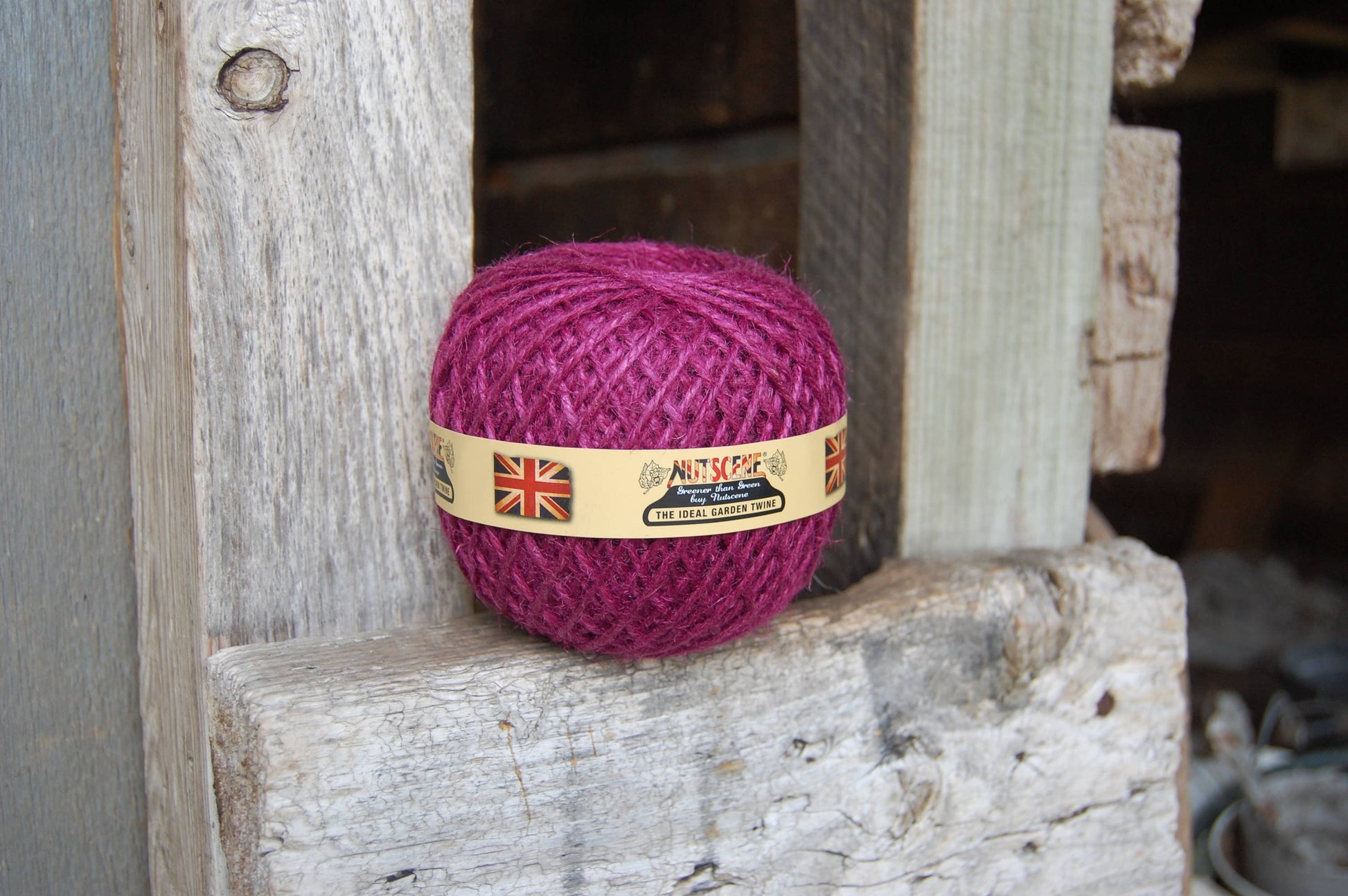 Ein magentafarbenes Knäuel des Heritage Twine von Nutscene Twines Ltd. sitzt auf verwitterten Holzbrettern, mit einem Etikett, das die britische Flagge und einen Text zeigt.