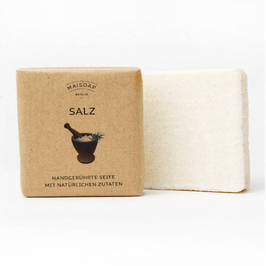 Ein quadratisches weißes Stück maisoap berlin - Salz Seife ist neben einer braunen Papierverpackung mit der Aufschrift "Salz" abgebildet, auf der ein Mörser und ein Stößel abgebildet sind und ein deutscher Text, der die Seife als handgemachte Gesichtsseife für trockene Haut mit natürlichen Inhaltsstoffen beschreibt.