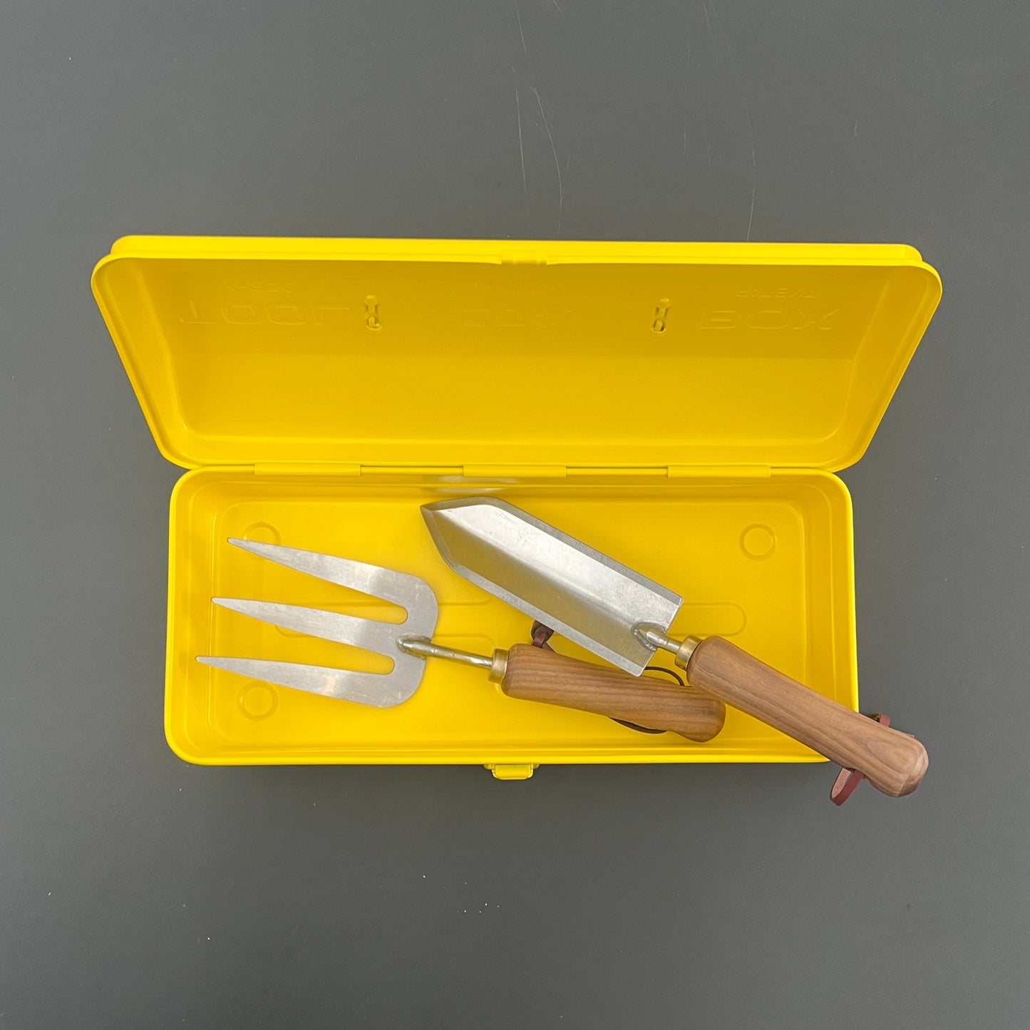 Eine gelbe Niwaki Y-Type Toolbox von Niwaki ist geöffnet und offenbart eine Handkelle aus Metall und eine dreizinkige Gabel mit Holzgriff - wesentliche Werkzeuge für den Gartenbedarf - auf grauem Hintergrund.