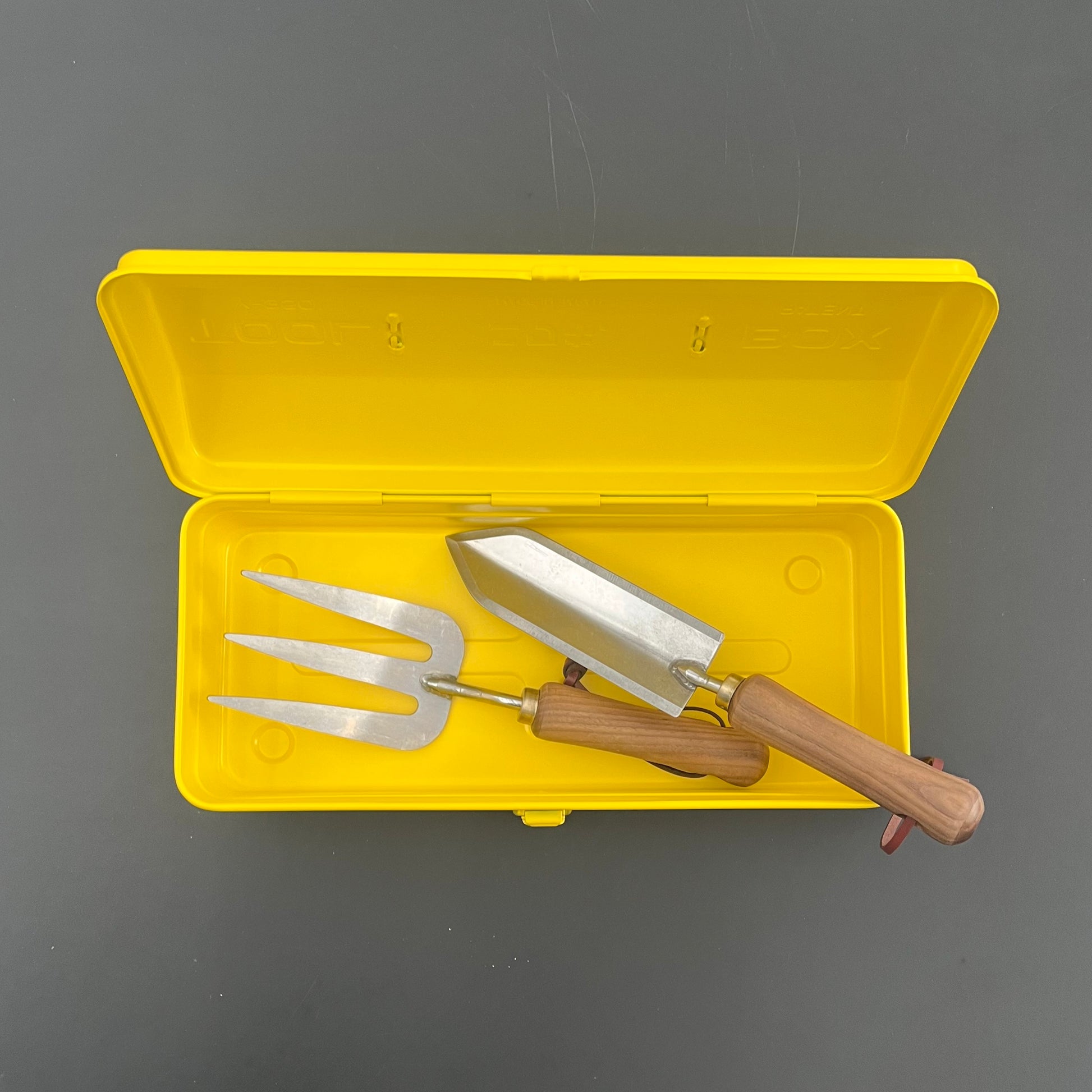 Eine gelbe Niwaki Y-Type Toolbox von Niwaki ist geöffnet und offenbart eine Handkelle aus Metall und eine dreizinkige Gabel mit Holzgriff - wesentliche Werkzeuge für den Gartenbedarf - auf grauem Hintergrund.