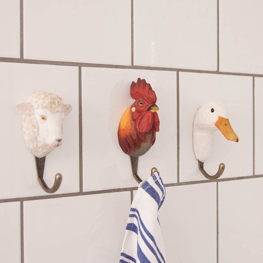 Der Handgeschnitzte Wandhaken aus Holz | Hahn von Wildlife Garden ist an einer weiß gekachelten Wand angebracht und hält ein blau-weiß gestreiftes Handtuch. Zwei weitere Tierhaken - ein Schaf und eine Ente - stehen daneben. Eine einzigartige Geschenkidee!.
