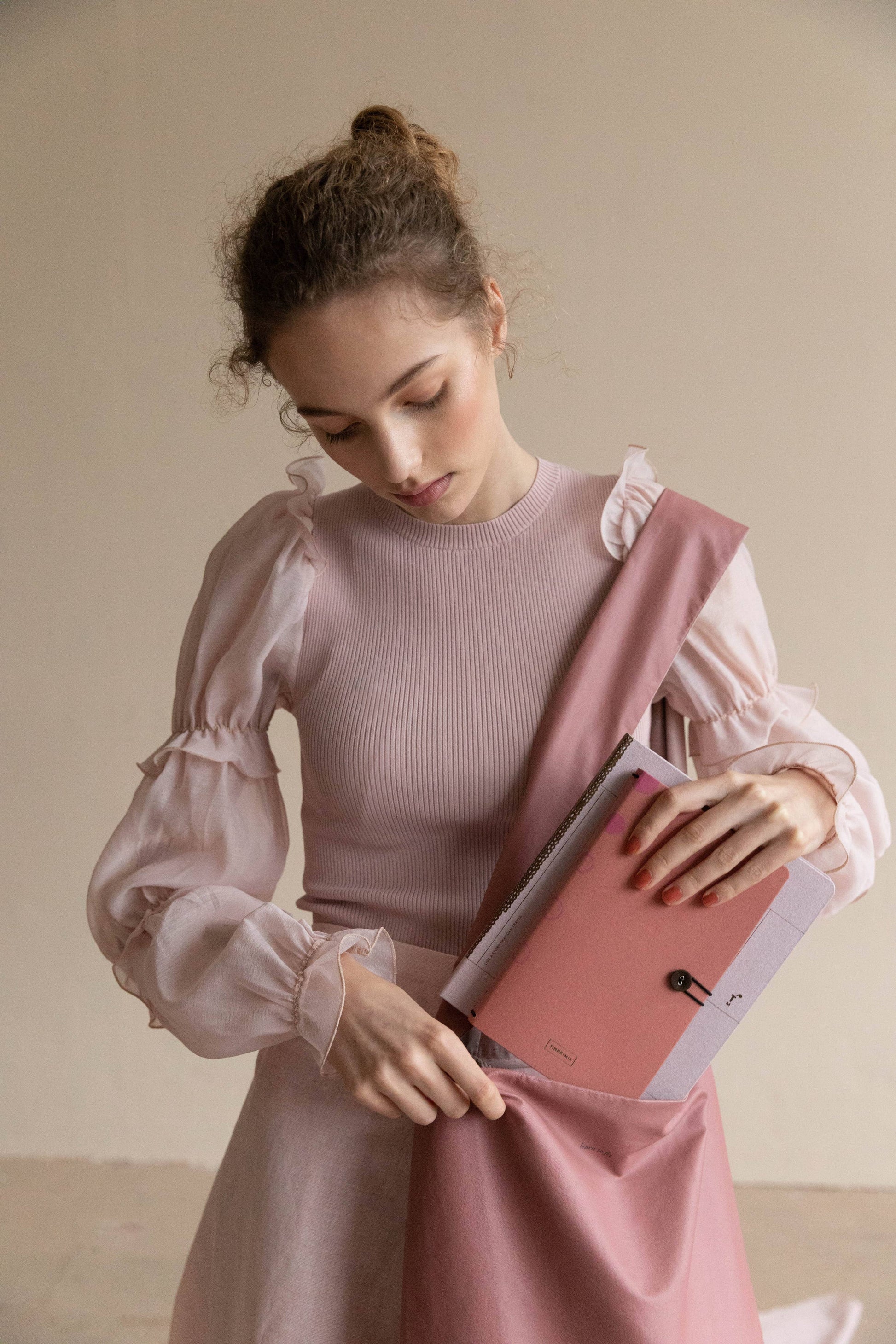 Eine junge Frau mit lockigem Haar in einer hellrosa Bluse legt das Tinne Mia Notizbuch mit Knopf - Leinen - Rose Dawn FSC Mix in eine passende rosa Tragetasche und blickt ruhig auf einen neutralen Hintergrund.