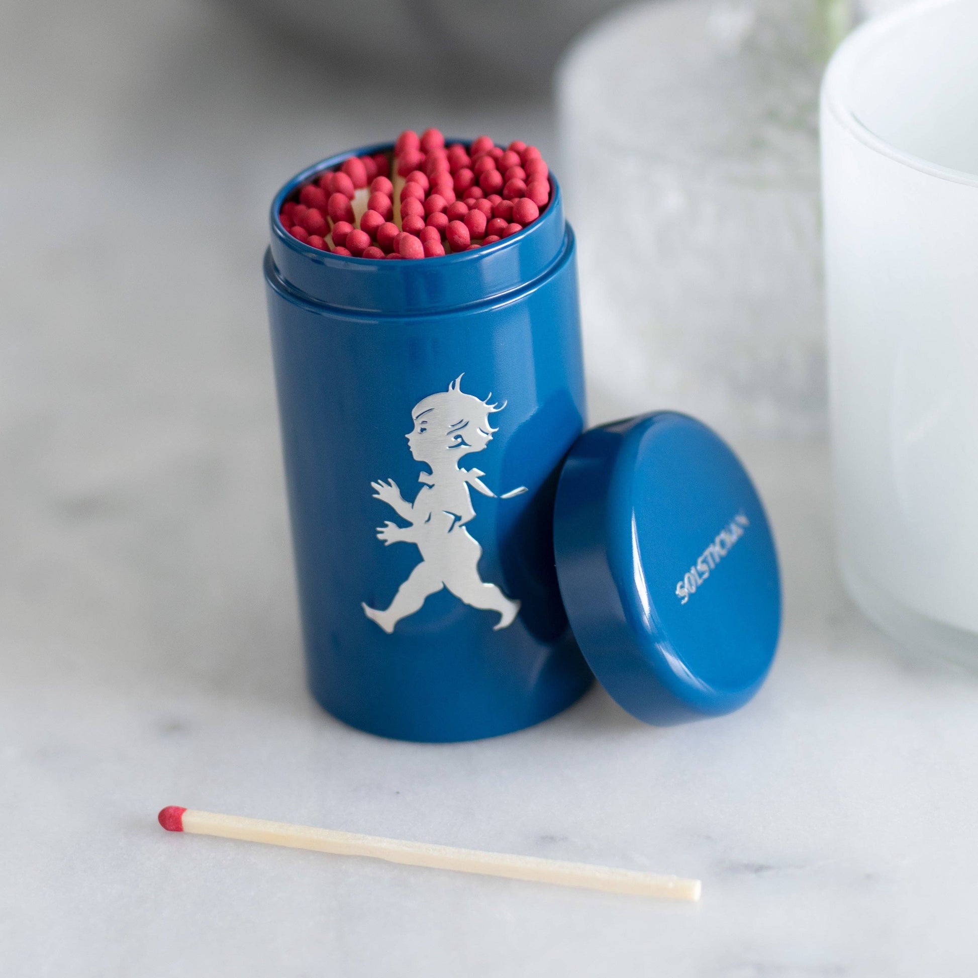 Ein blauer Solstickan Design Passender Schlauch (nur EU) mit weißer Solsticke Boy-Illustration fasst rote Streichhölzer, der Deckel liegt daneben und ein Streichholz davor auf einer weißen Fläche.