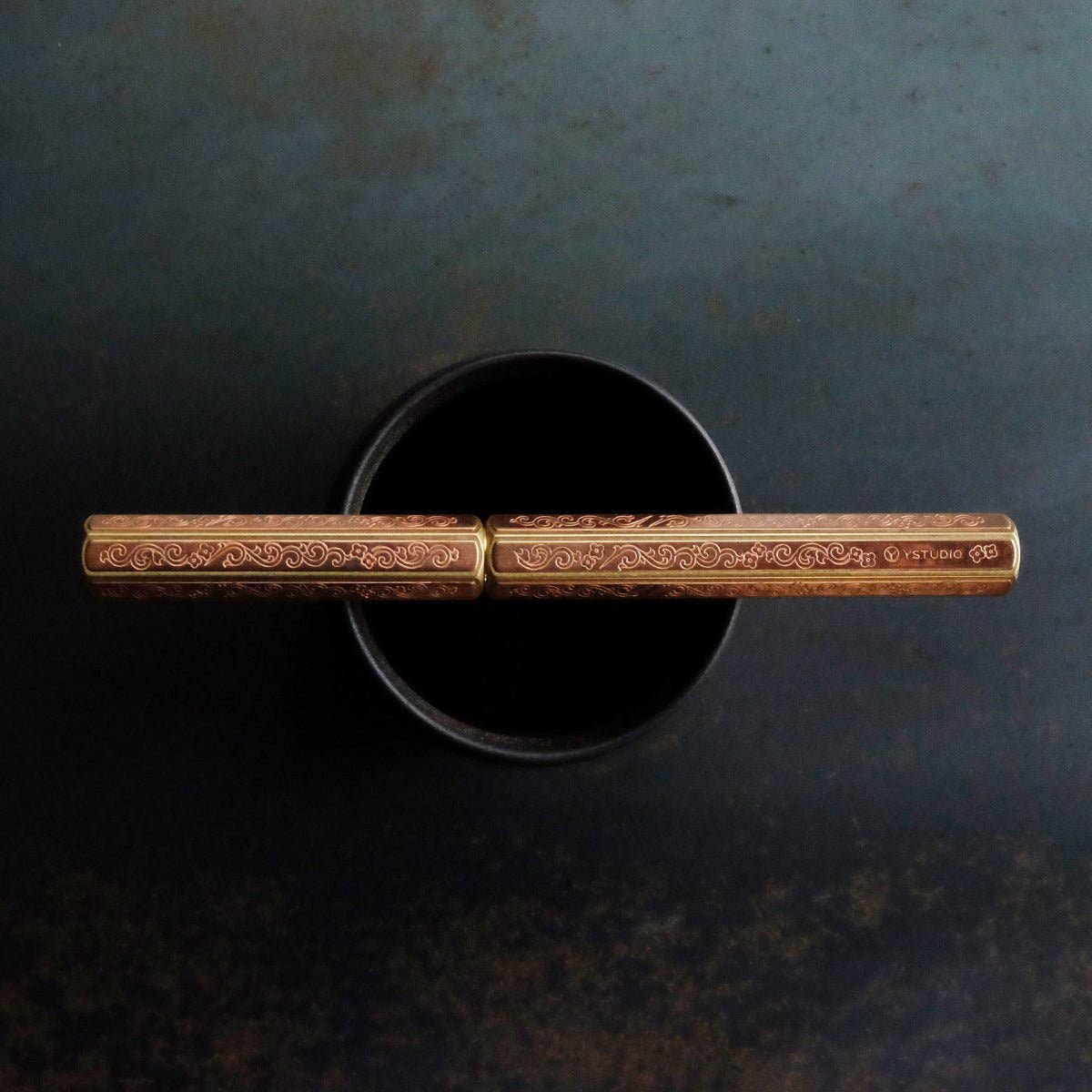 Ein Paar goldener Essstäbchen, inspiriert von der Metallgravurkunst, in Anlehnung an die Designs von Kazari Kanagu, ruht auf einer dunklen Schale. Inspiriert von ENTREPÔTs YSTUDIO Klassische Renaissance KAZARI KANAGU ORCHID Tintenroller.