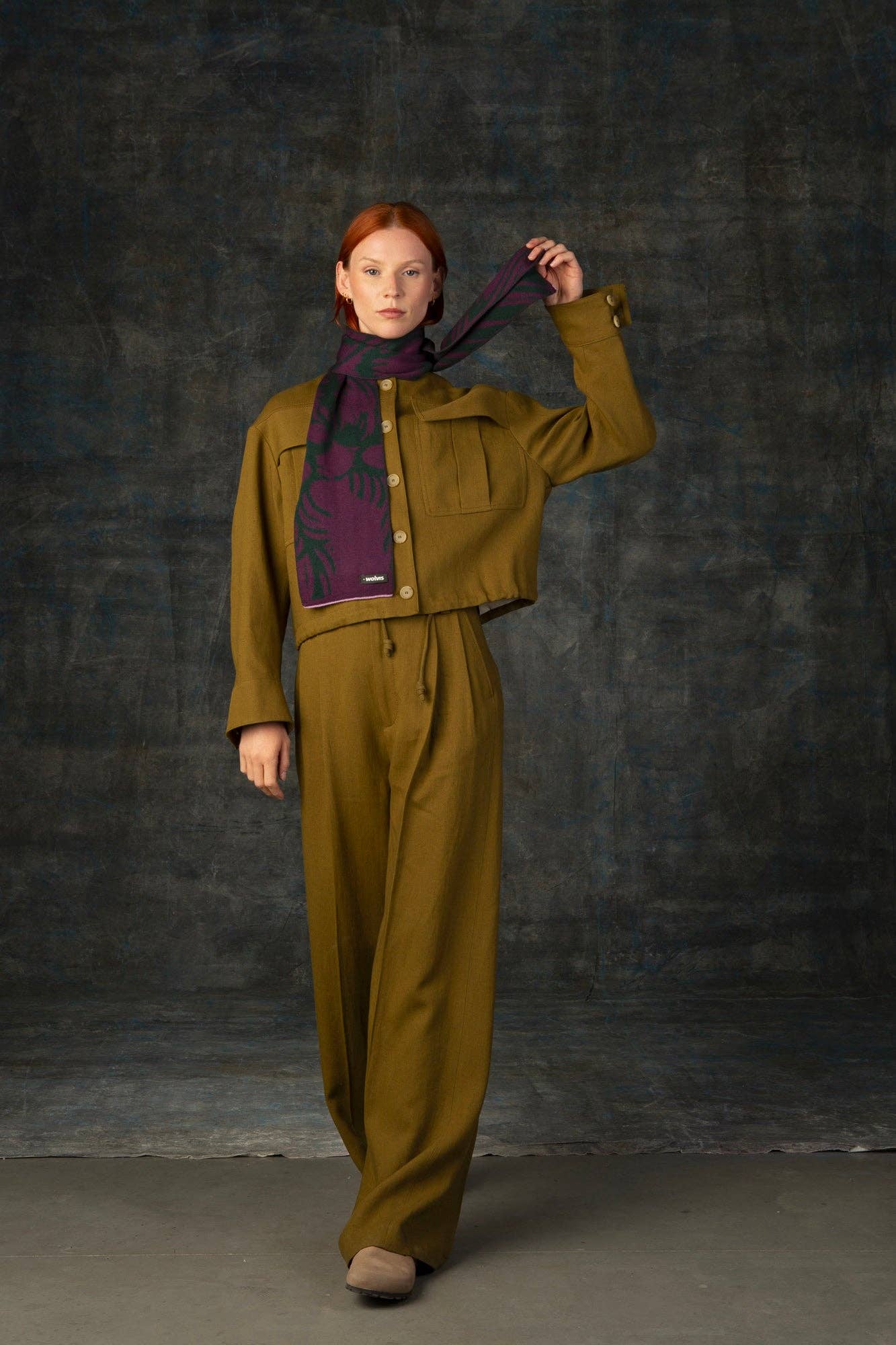 Eine rothaarige Frau in einem olivgrünen Hosenanzug und beigen Schuhen steht vor einem dunkel strukturierten Hintergrund und hält den Wolvis Schal aus Merinowolle - Cerura Traube Mini mit seinem eleganten Jacquardmuster um den Hals.