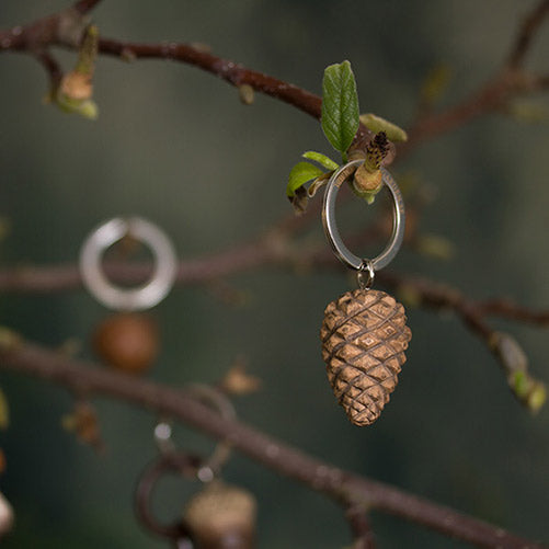 Ein handgefertigter Schlüsselanhänger aus Holz | Zapfen von Wildlife Garden hängt an einem Zweig mit frischen grünen Blättern, im Hintergrund ist ein weiterer unscharfer Anhänger erkennbar.
