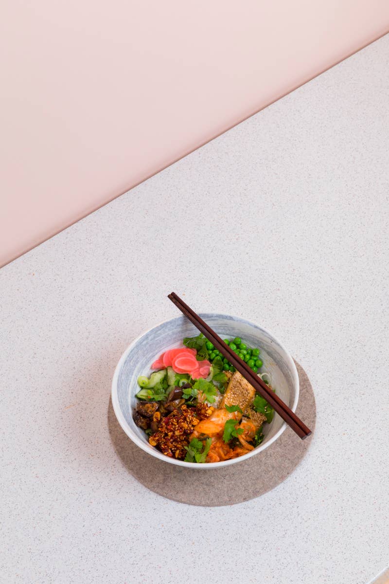 Eine Schale mit lebhaften asiatischen Nudeln mit Grünzeug, eingelegtem Gemüse, Chili-Öl und frischen Kräutern wird mit der Erdnuss-Rayu-Soße von White Mausu Ltd. serviert. Auf einer runden Matte platziert, liegen Stäbchen daneben und runden das Ganze ab.