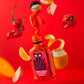 Eine Flasche Crazy Bastard Sauce - Trinidad Scorpion & Clementine 100 ml von CBS Foods schwebt in der Luft, ohne Deckel, die Sauce spritzt heraus, umgeben von Apfelscheiben, Orangenschalen und rotem Paprika vor einem leuchtend roten Hintergrund.