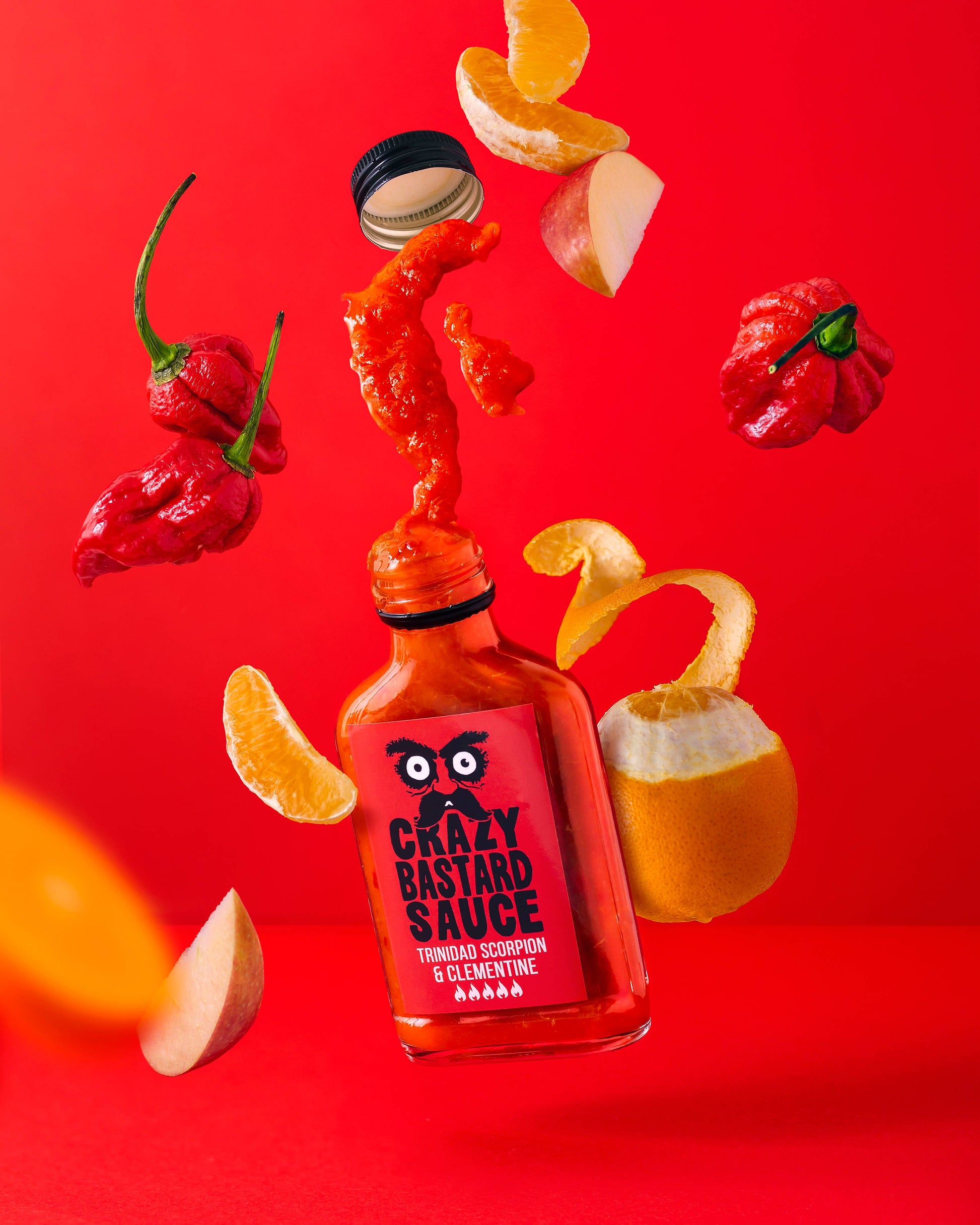 Eine 100-ml-Flasche der CBS Foods GmbH (Crazy Bastard Sauce) Trinidad Scorpion & Clementine scharfe Soße mit spritzender Soße, schwimmenden Clementinenschalen, Apfelscheiben und feurigen Trinidad Scorpion-Schoten auf einem leuchtend roten Hintergrund.