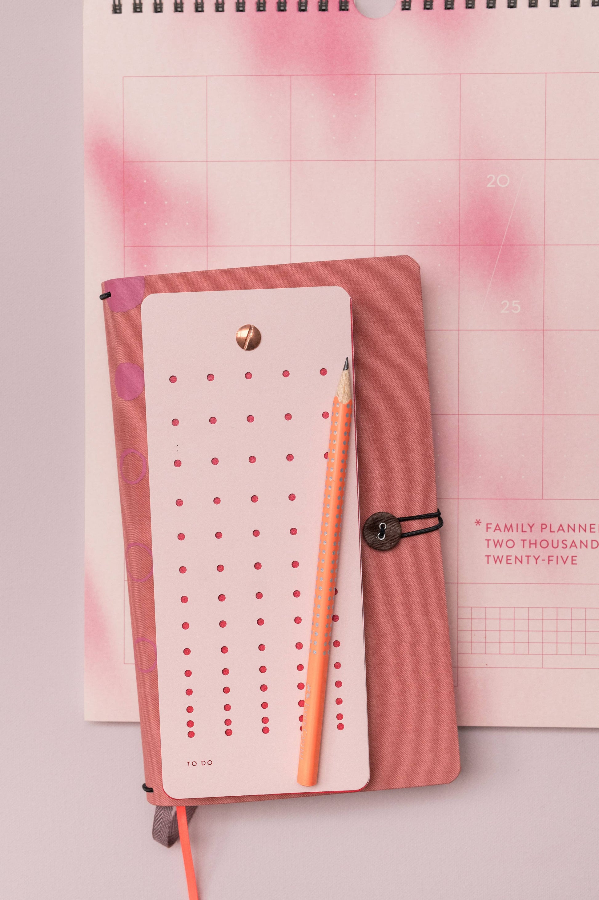 Ein pinkfarbenes Flatlay zeigt die Tinne Mia - Aufgabenliste - Mellow Rose, einen 2025 Familienplaner-Kalender und ein pinkfarbenes Listenbuch mit einem orangefarbenen Stift - alles sauber arrangiert auf FSC-zertifiziertem Papier für einen nachhaltigen Touch.