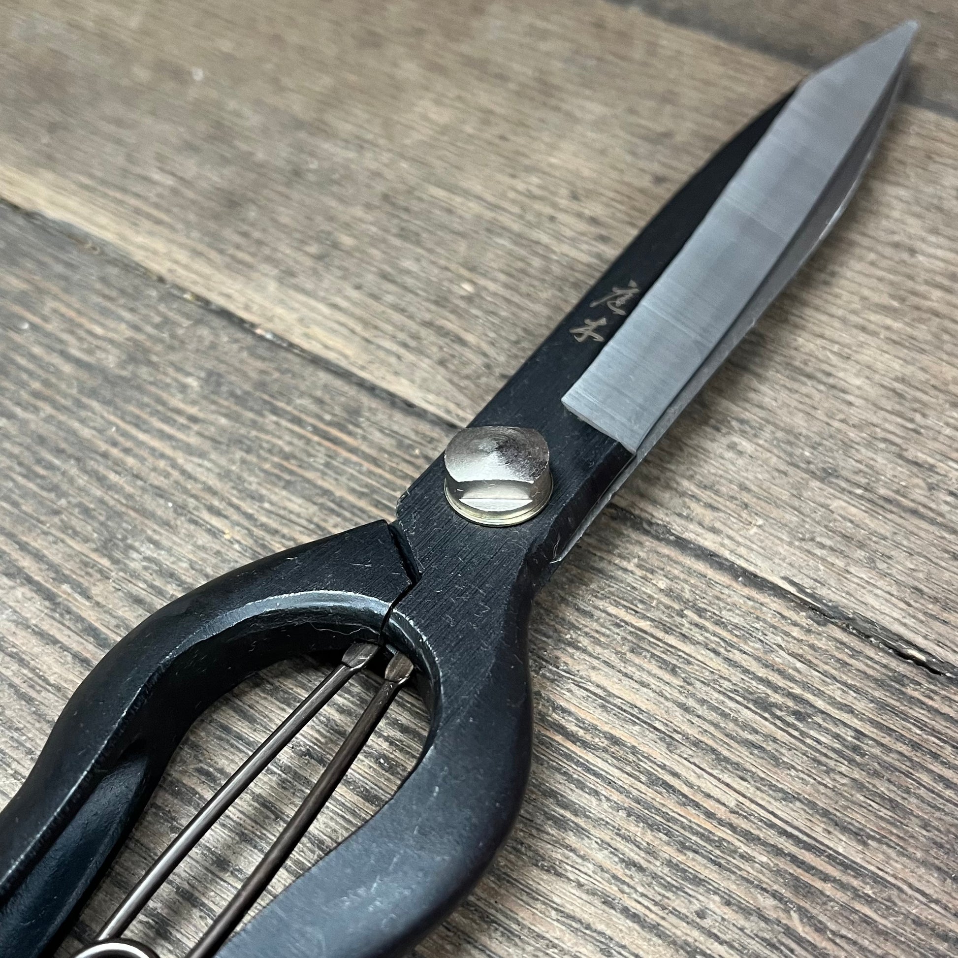 Nahaufnahme der Niwaki Sentei Topiary Clippers von Niwaki, mit einer scharfen, spitzen schwarzen Klinge mit sichtbarer Metallschraube und Federmechanismus, die auf einer Holzoberfläche ruht.