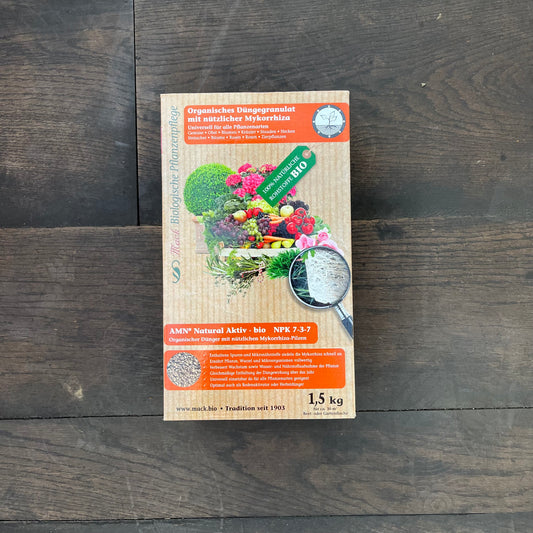 Auf einem dunklen Holzboden steht eine 1,5 kg schwere Schachtel mit dem organischen Dünger AMN Natural Aktiv-bio von AMN, der natürliche Mykorrhiza enthält und auf dessen Etikett Bilder von Gemüse und Gartenprodukten zu sehen sind.