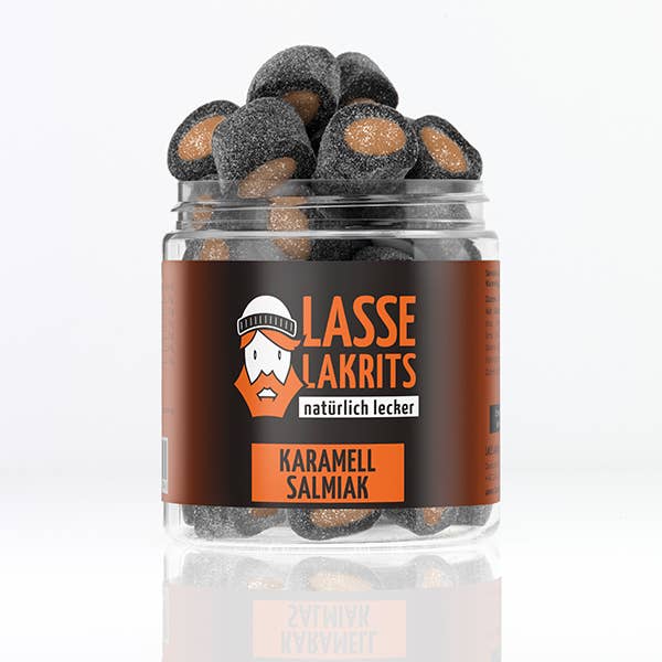 Die LASSE LAKRITS - natürlich lecker - Dose Karamell Salmiak aus durchsichtigem Kunststoff ist mit schwarzen, gezuckerten Bonbons mit Karamellkern gefüllt. Das schwarze Etikett zeigt "LASSE LAKRITS" in Orange und Weiß sowie ein bärtiges Cartoon-Gesicht.