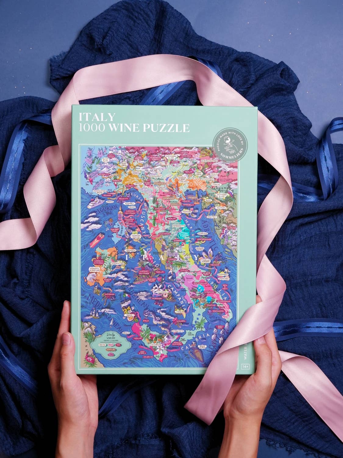 Eine Person hält das Water & Wines - Wein Puzzle - Italien, ein farbenfrohes 1000-teiliges Puzzle von Water & Wines mit einer illustrierten Karte der italienischen Weinregionen und Rebsorten, vor einem blauen Stoff mit einem rosa Band.