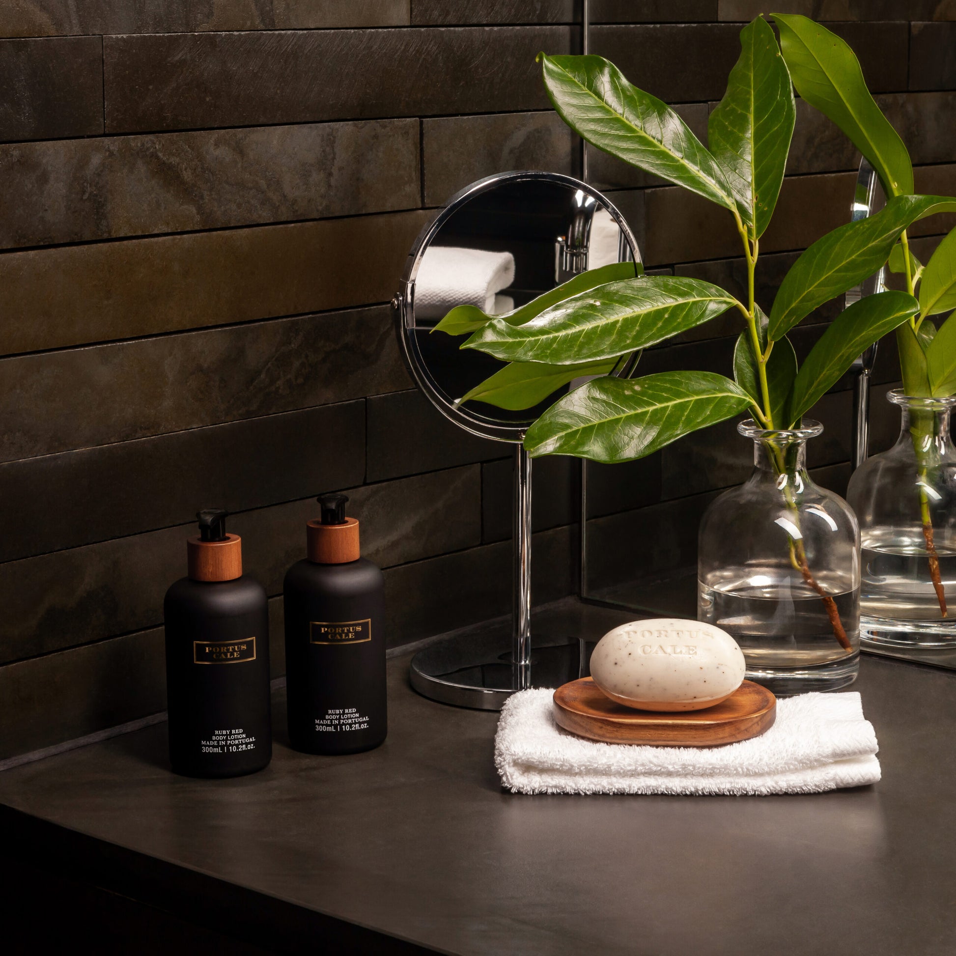 Ein moderner Badezimmertisch mit zwei schwarzen Seifenspendern, einem runden Spiegel, einer Glasvase mit grünen Blättern und der Castelbel's Body Lotion Portus Cale 300 ml - Ruby Red auf einem weißen Handtuch auf einem Holztablett, alles vor dunkel gefliesten Wänden.