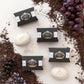 Fünf schwarze Schachteln Castelbel Seife Portus Cale 40 g - Ruby Red und drei weiße Pflanzenseifen liegen auf einer weißen Fläche, umgeben von losen Teeblättern und roten und schwarzen Weintrauben.