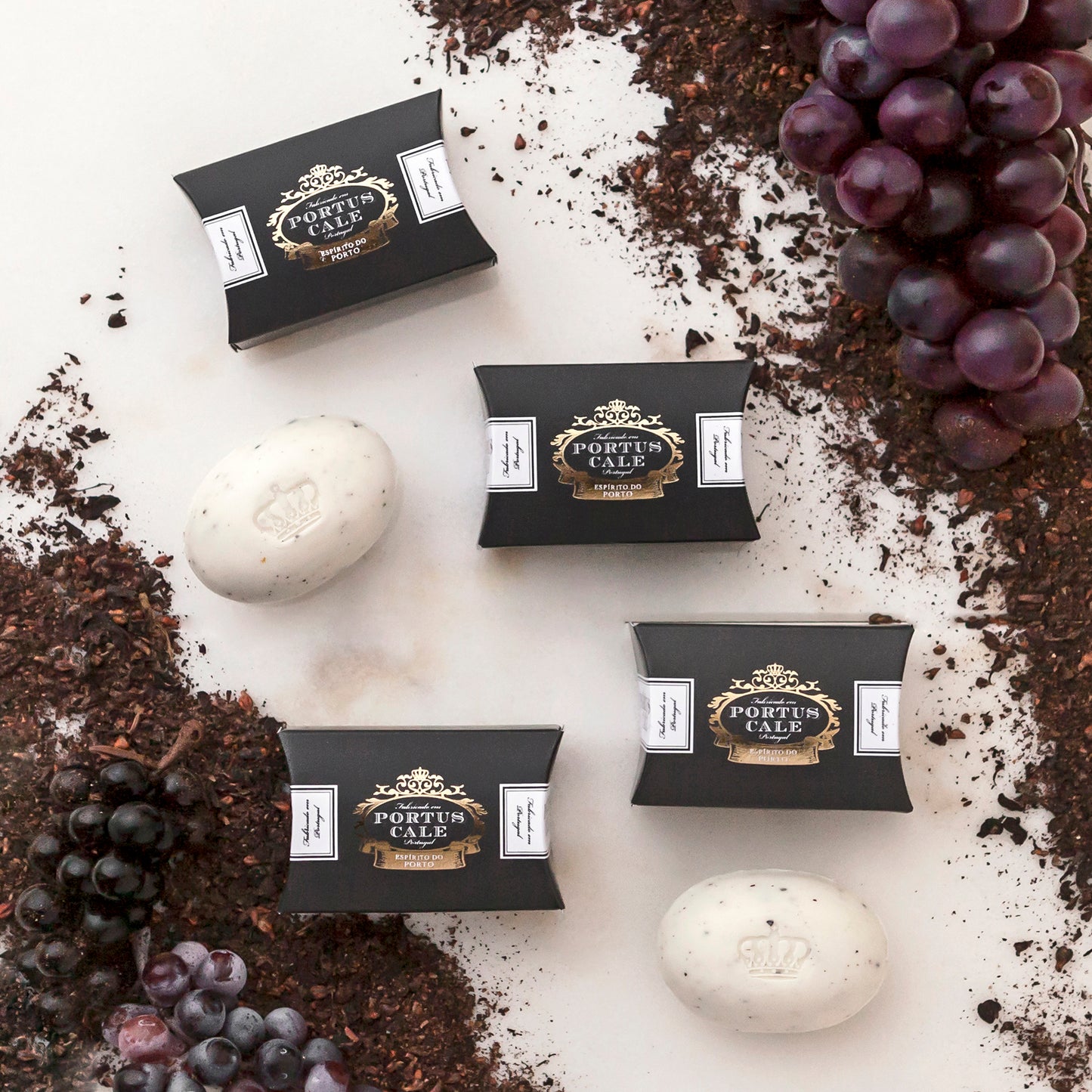 Fünf schwarze Schachteln Castelbel Seife Portus Cale 40 g - Ruby Red und drei weiße Pflanzenseifen liegen auf einer weißen Fläche, umgeben von losen Teeblättern und roten und schwarzen Weintrauben.