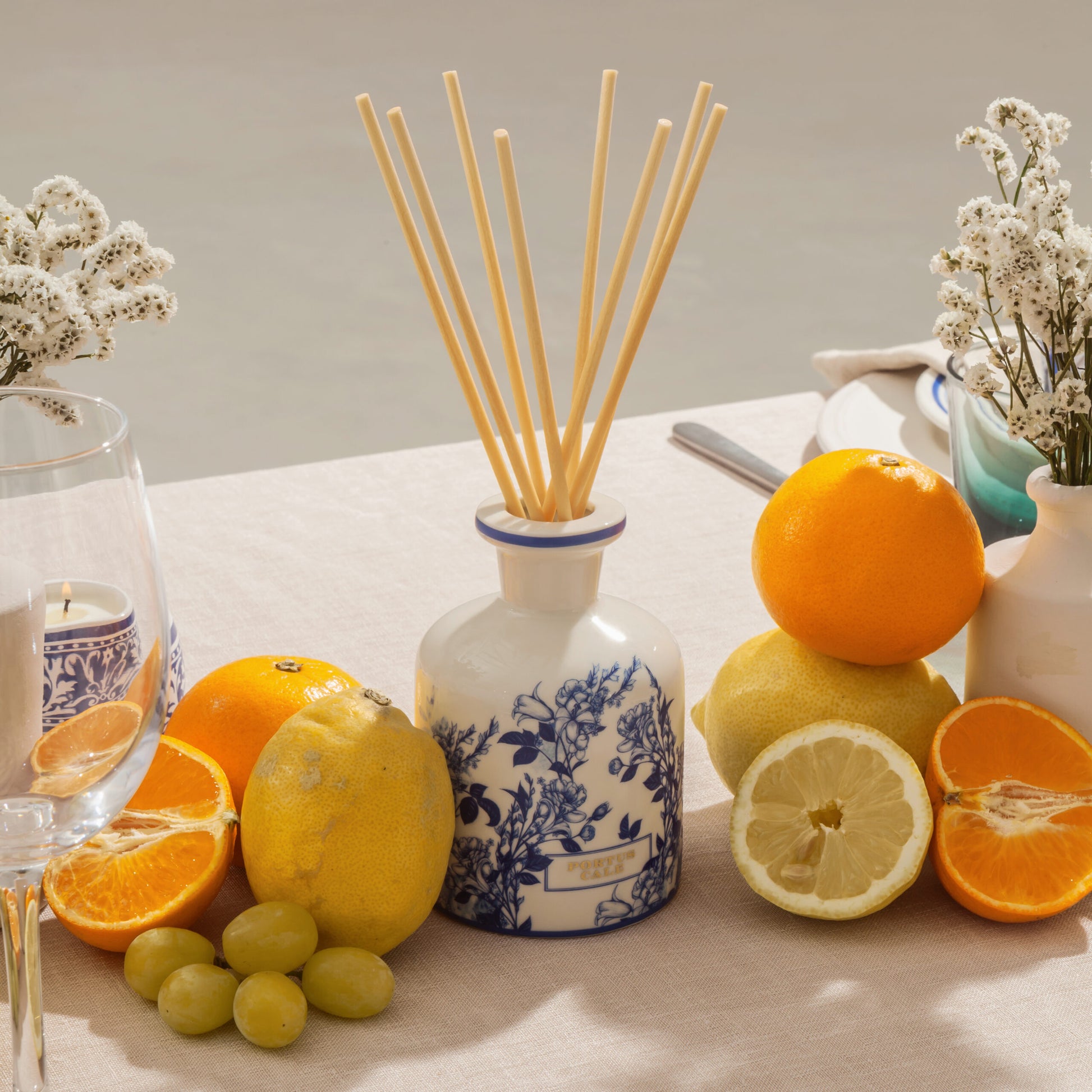 Ein Tisch mit einem Castelbel Diffusor | Gold & Blue 250 ml, umgeben von Orangen, Zitronen, Trauben, weißen Blumen in Vasen, einem Glas und einem Teller auf einer beigen Tischdecke.