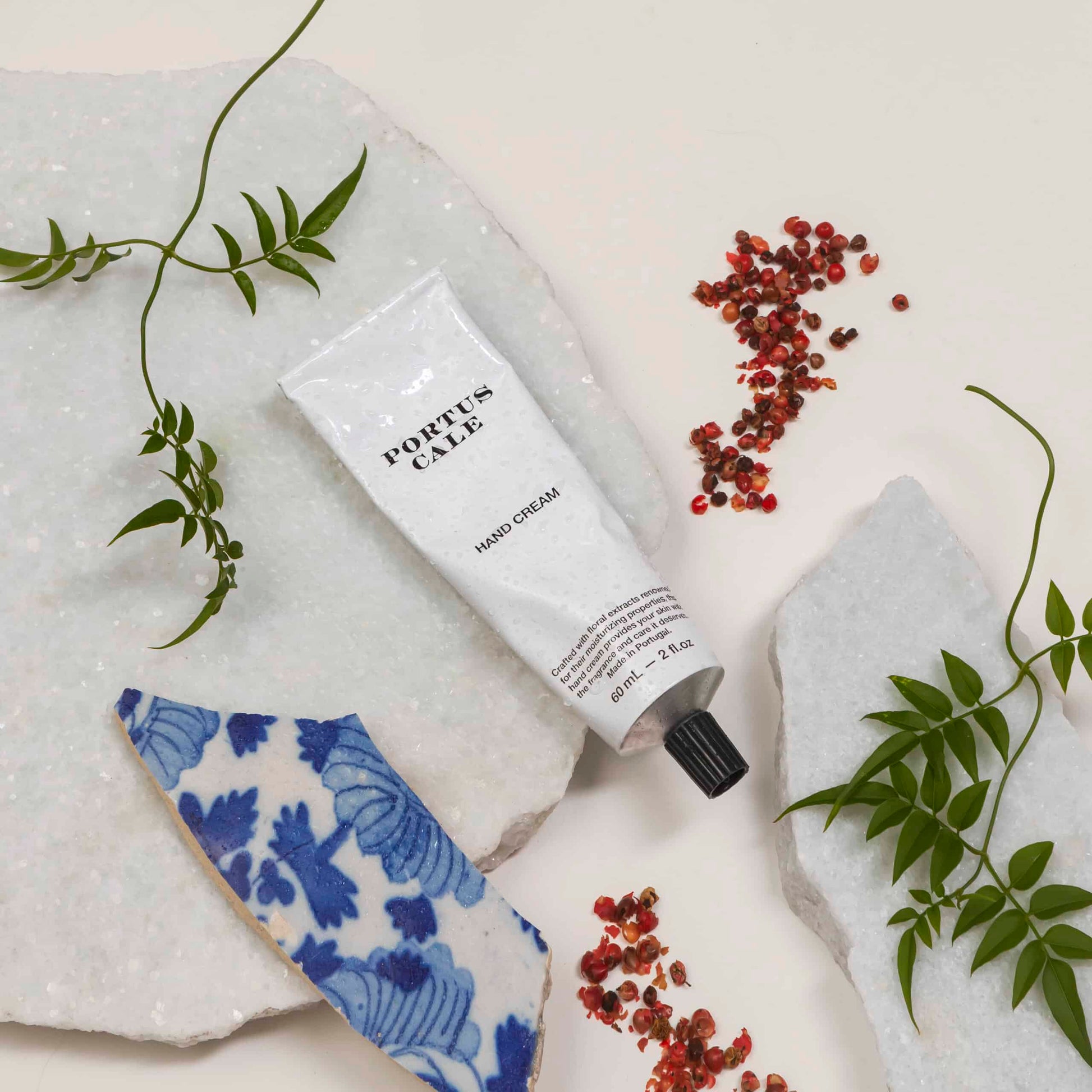 Eine Tube Castelbel Handcreme | Gold & Blau 60 ml ruht auf einer weißen Marmorfläche, umgeben von grünen Blättern, roten Samen und einem blau-weiß gemusterten Keramikstück.