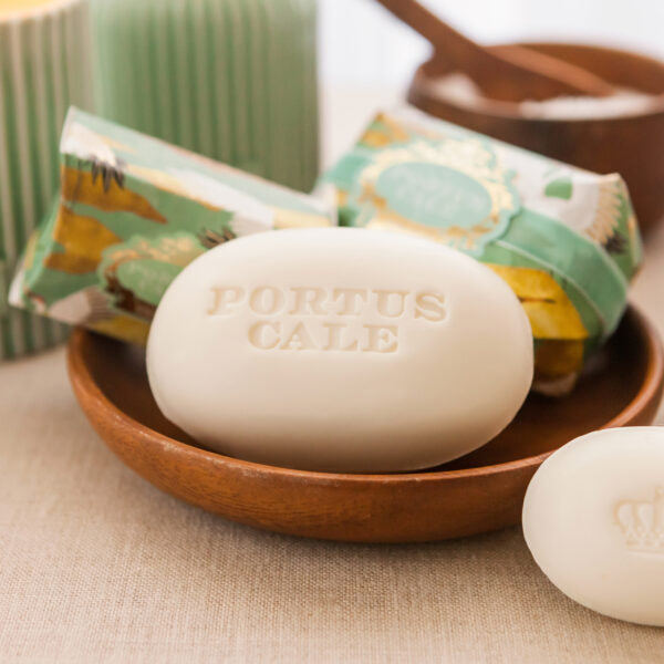 Ein weißes Stück Castelbel Seife | White Crane 150 g, hergestellt aus pflanzlichen Inhaltsstoffen und mit der Prägung PORTUS CALE, liegt in einer Holzschale. Nebenan sorgen eine verpackte Seife und ein weiteres Stück für ein sanftes, natürliches Ambiente.