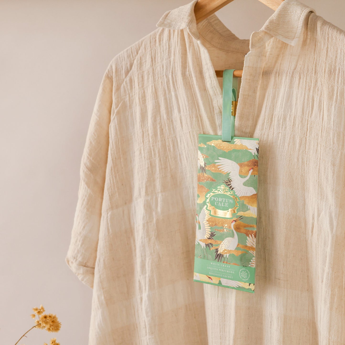 Ein beigefarbenes, strukturiertes Kleid hängt an einem Holzbügel, geschmückt mit einem Castelbel White Crane Sachet (10g). Das Etikett zeigt weiße Kraniche und grünes Laub. Der Duft von Yuzu, Vetiver und Mandarine erfüllt die Luft, während gelbe Blumen die Ecke erhellen.