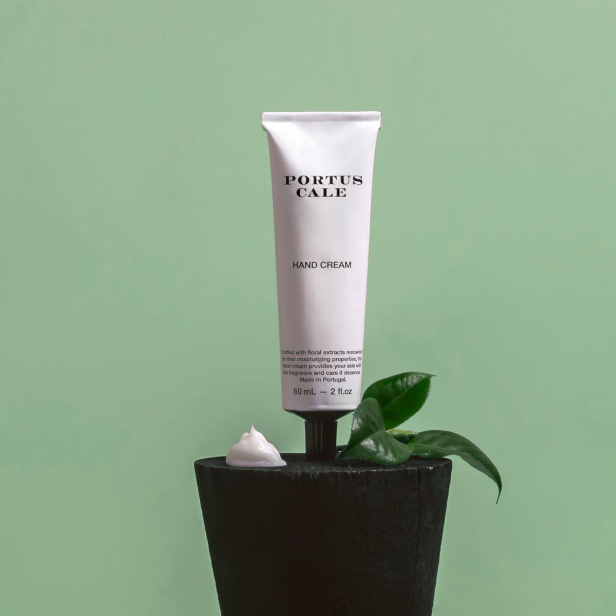 Eine Tube Castelbel Handcreme | Weißer Kranich 60 ml steht aufrecht auf einem schwarzen Topf mit grünen Blättern und einem Klecks Handcreme davor, vor einem hellgrünen Hintergrund.