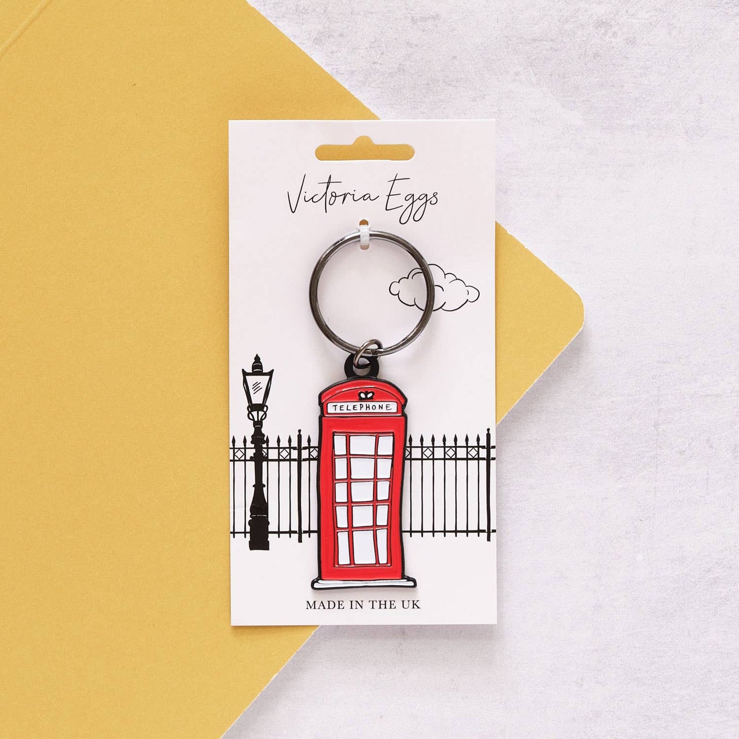 Der Victoria Eggs - Telefonzelle Schlüsselanhänger, inspiriert von der britischen Ikone, hängt an einer Victoria Eggs Karte mit schwarzem Zaun und Laternenpfahl – ein charmantes Geschenk für Fans britischer Kultur.