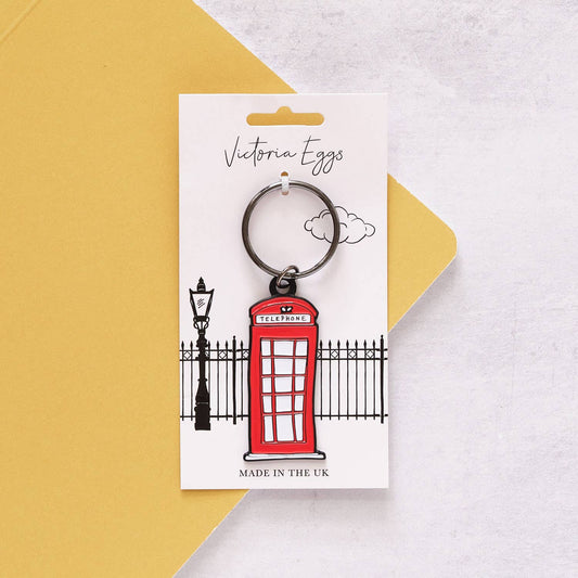 Der Victoria Eggs - Telefonzelle Schlüsselanhänger, inspiriert von der britischen Ikone, hängt an einer Victoria Eggs Karte mit schwarzem Zaun und Laternenpfahl – ein charmantes Geschenk für Fans britischer Kultur.