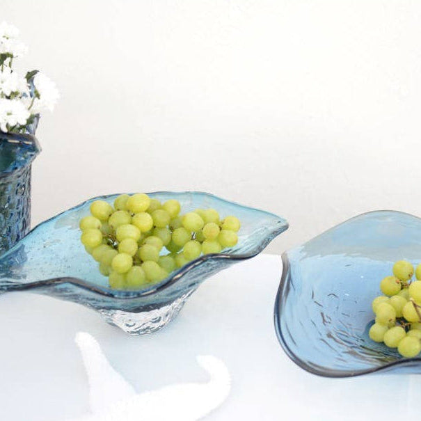 Zwei AULICA - Dekorative blaue Glasschale 33x18x10,5cm halten grüne Weintrauben auf einer weißen Oberfläche. Eine blaue AULICA Vase mit weißen Blumen und einem Seestern ergänzen dieses leichte, luftige Tischset.