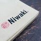 Eine beige Stofftasche für Gärtnerzubehör von Niwaki mit „Niwaki“ in schwarzer Schrift und einem roten, kreisförmigen Logo im japanischen Stil liegt auf einer dunklen Holzoberfläche – perfekt für die Gartenarbeit oder zur Aufbewahrung Ihres Niwaki Kniekissens.