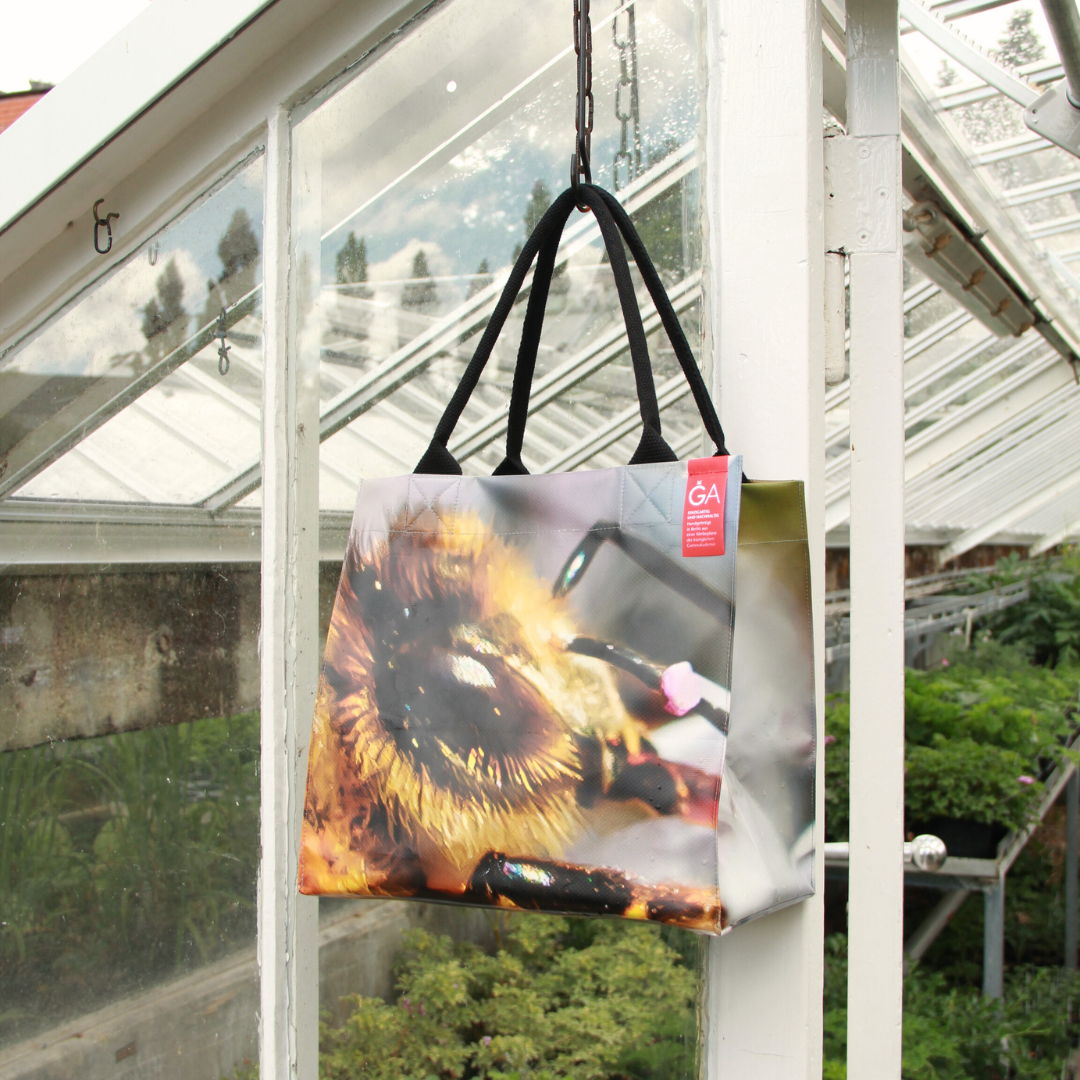 Die KGA Upcycling KGA Tasche 'Biene', eine Unikat-Tasche mit schwarzen Henkeln und einem Bienenmotiv in Nahaufnahme, hängt an einem Haken in einem gläsernen Gewächshaus, aus dem man Pflanzen sehen kann.