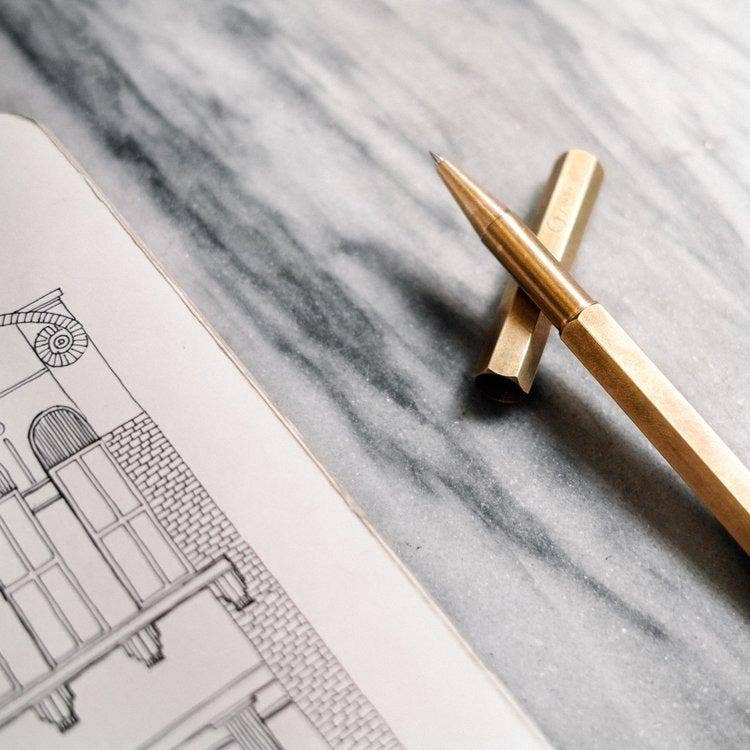 Der ENTREPÔT - YSTUDIO Klassischer Revolve-Rollerball-Stift aus Messing von ENTREPÔT liegt auf grauem Marmor neben einem offenen Notizbuch mit architektonischer Zeichnung - perfekt für sanftes Schreiben.